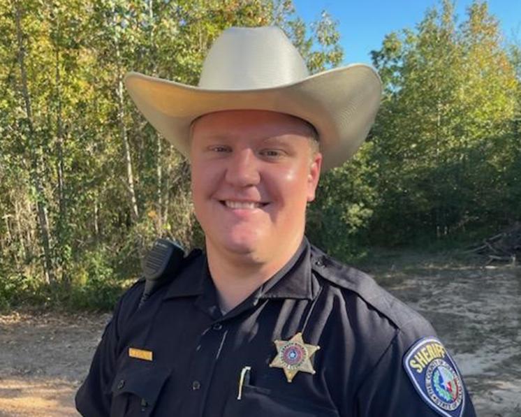 A man smiling at the camera in sheriff clothes and a cowboy hat on with nature in the background
