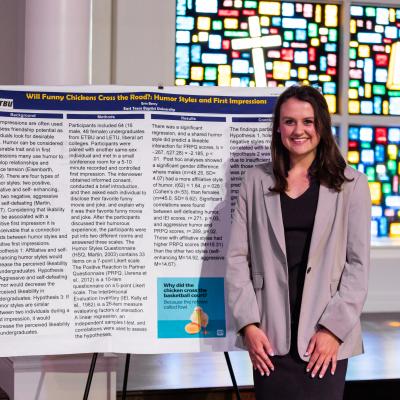 Student stands with research project poster board in front of a stained glass window