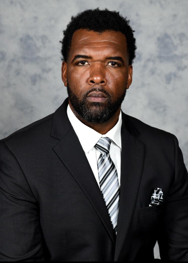 A headshot of a man in front of grey background in a black suit.