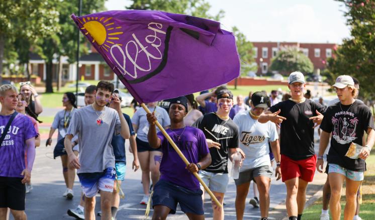 fall_2021_first_week_img_3 A group of people walking outdoors, with one person holding a large purple flag that has a yellow sun and the word "Rebels."