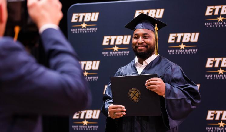 2022_commencement_fall_3 A man smiling at a camera in his graduation regalia holding his diploma up with a navy backdrop