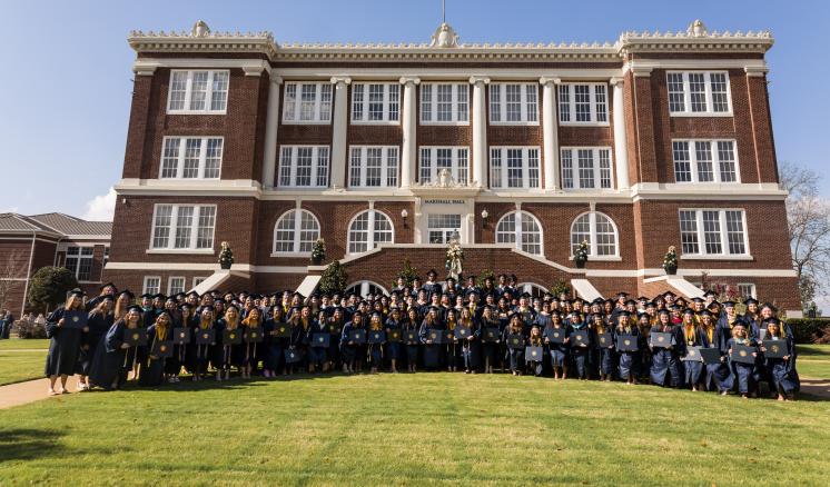 Large group of graduates posed with diplomas in front of brick academic building smiling at the camera