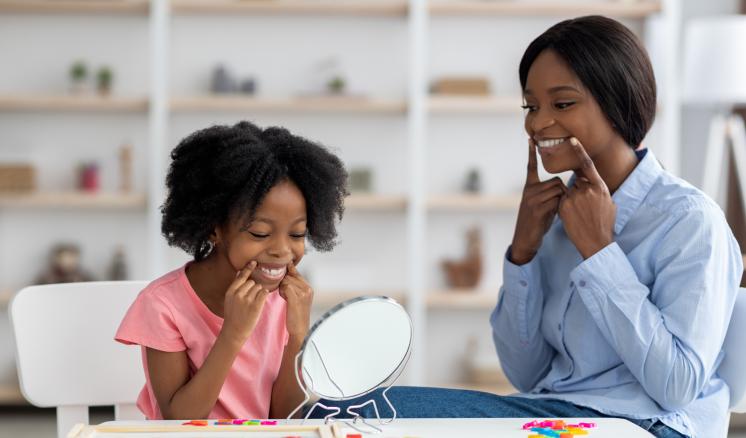 MS-SLP Leveling Track A child and speech-language pathologist sit together at a table, both smiling warmly and pointing to their cheeks in front of a small mirror.