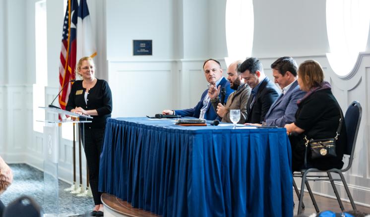 A group of people seated at a table with a blue tablecloth, while a public speaker stands at the podium in front. Formal event, inside