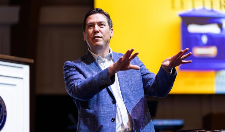 A man is standing on a stage with a projector screen behind him, using his hands to add movement to his public speech