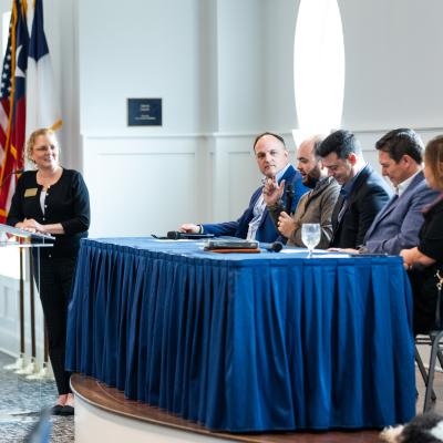 A group of people seated at a table with a blue tablecloth, while a public speaker stands at the podium in front. Formal event, inside