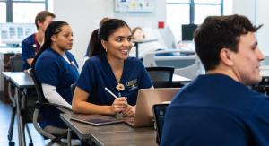 Students in blue scrubs seated in a classroom with laptops, medical training mannequins in the background.