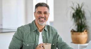 A man sitting at a table holdiong a coffee cup with papers on the table, in a brightly lit room, with a window behind him