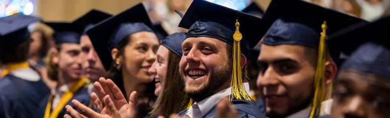 img_9 Graduates in navy caps and gowns clapping during a ceremony.