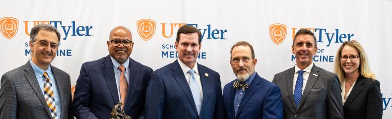 UT Tyler School of Medicine EAP Group of five males and one female smiling at camera in front of a white UT Tyler backdrop