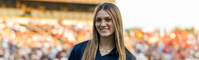 Haylie Stum A female student holding a microphone on a football field with the stands full of fans in the background.