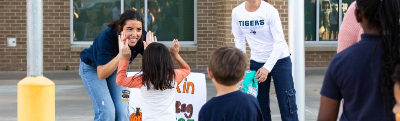 2024_misd_fall_festival_0584 Two volunteers engage with children at a fall festival activity, sharing high-fives in front of a sign that reads "Pumpkin Bag Toss."